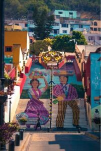 Imagen de una escalinata pintada con la figura de un hombre y una mujer en trajes típicos con un reloj de flores en la parte más alta, en Atlixco, Puebla.